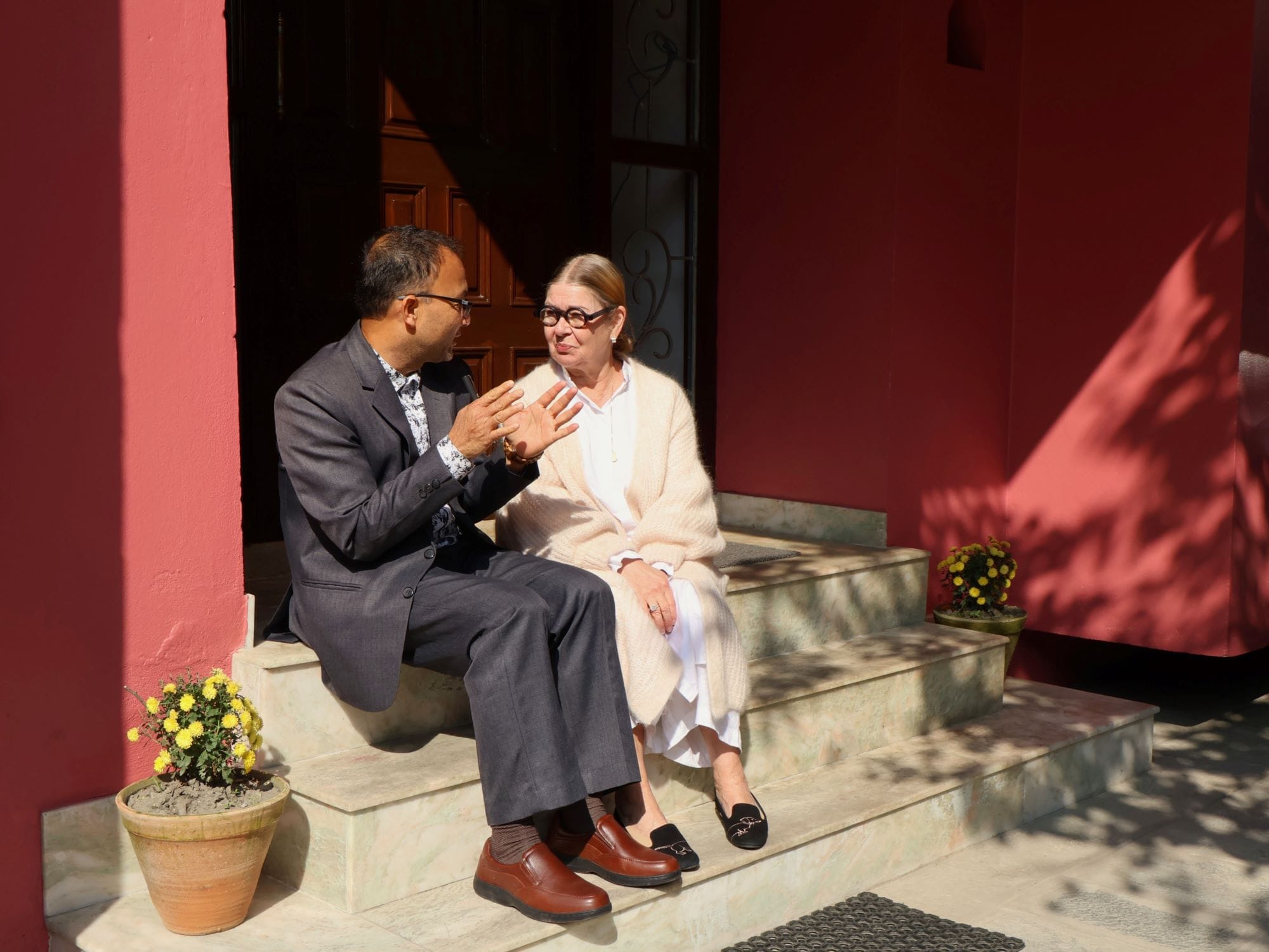 Two people sitting on outdoor steps, engaged in conversation. Both are dressed in formal attire. The background features a red wall and potted plants.